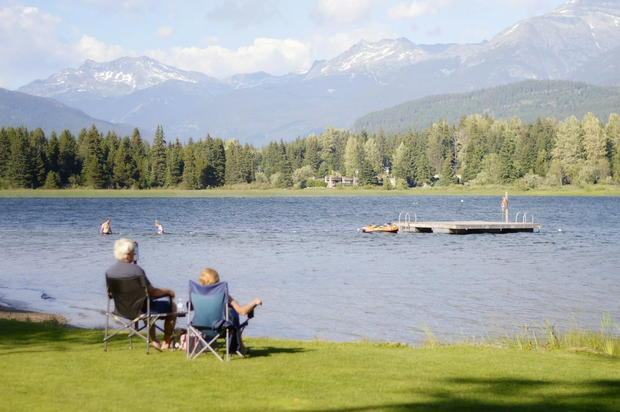 A retired couple in camping chairs on a lakeshore lawn, watching Alta Lake and the mountains beyond, in Whistler, British Columbia.