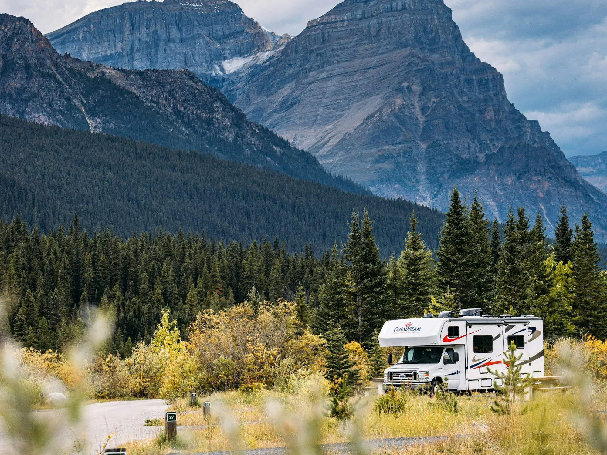 A motorhome parked at a campsite among autumn shrubs and spruce, below steep grey peaks on the Icefields Parkway, Alberta.