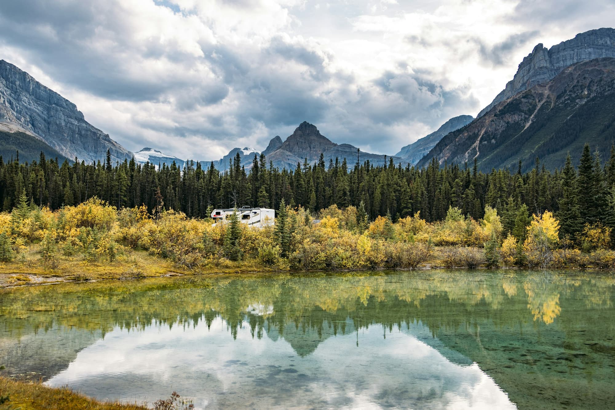 A white RV parked among autumn-gold shrubs beside a still mountain lake, with spruce forest and rugged peaks beyond.