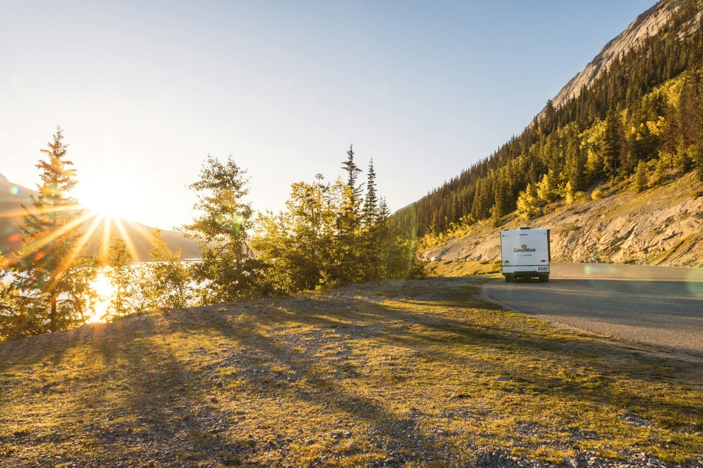 An RV driving a mountain highway at sunset, sunlight flaring through the trees.