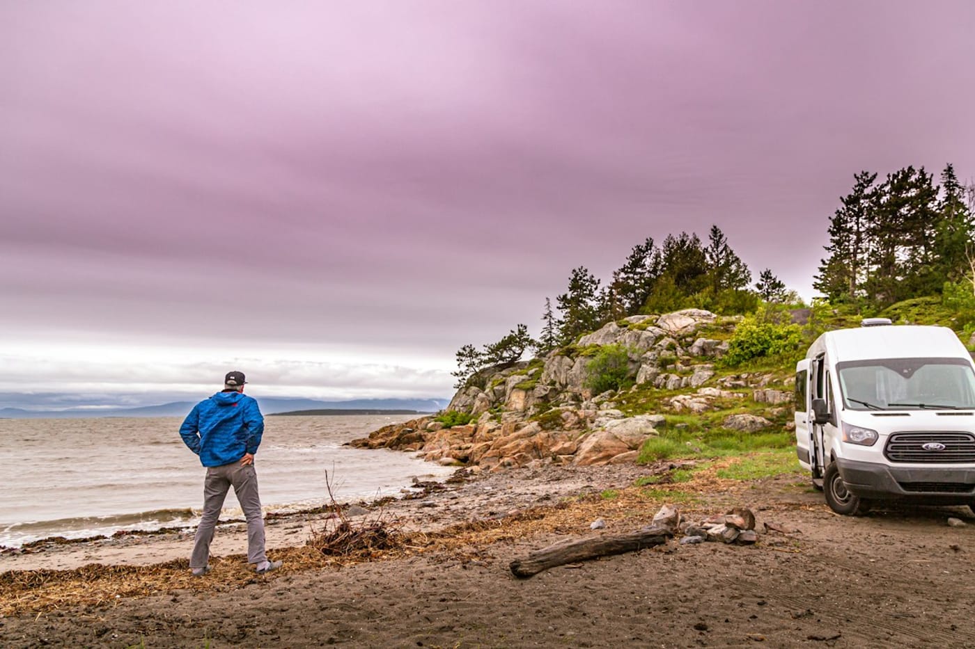A person in a blue jacket stands on a shoreline beside a parked white van under a pale pink dusk sky.