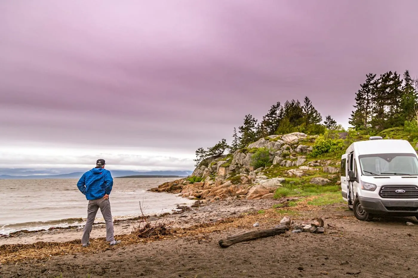 A person in a blue jacket stands on a shoreline beside a parked white van under a pale pink dusk sky.