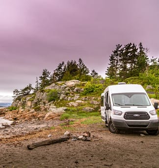 A person in a blue jacket stands on a shoreline beside a parked white van under a pale pink dusk sky.