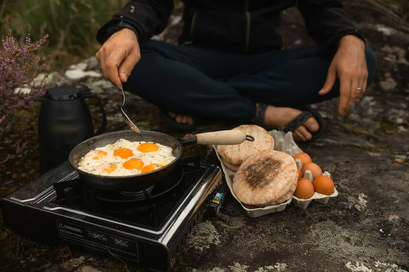 Fried eggs in a pan on a single-burner camp stove, with flatbreads and a carton of eggs beside it.