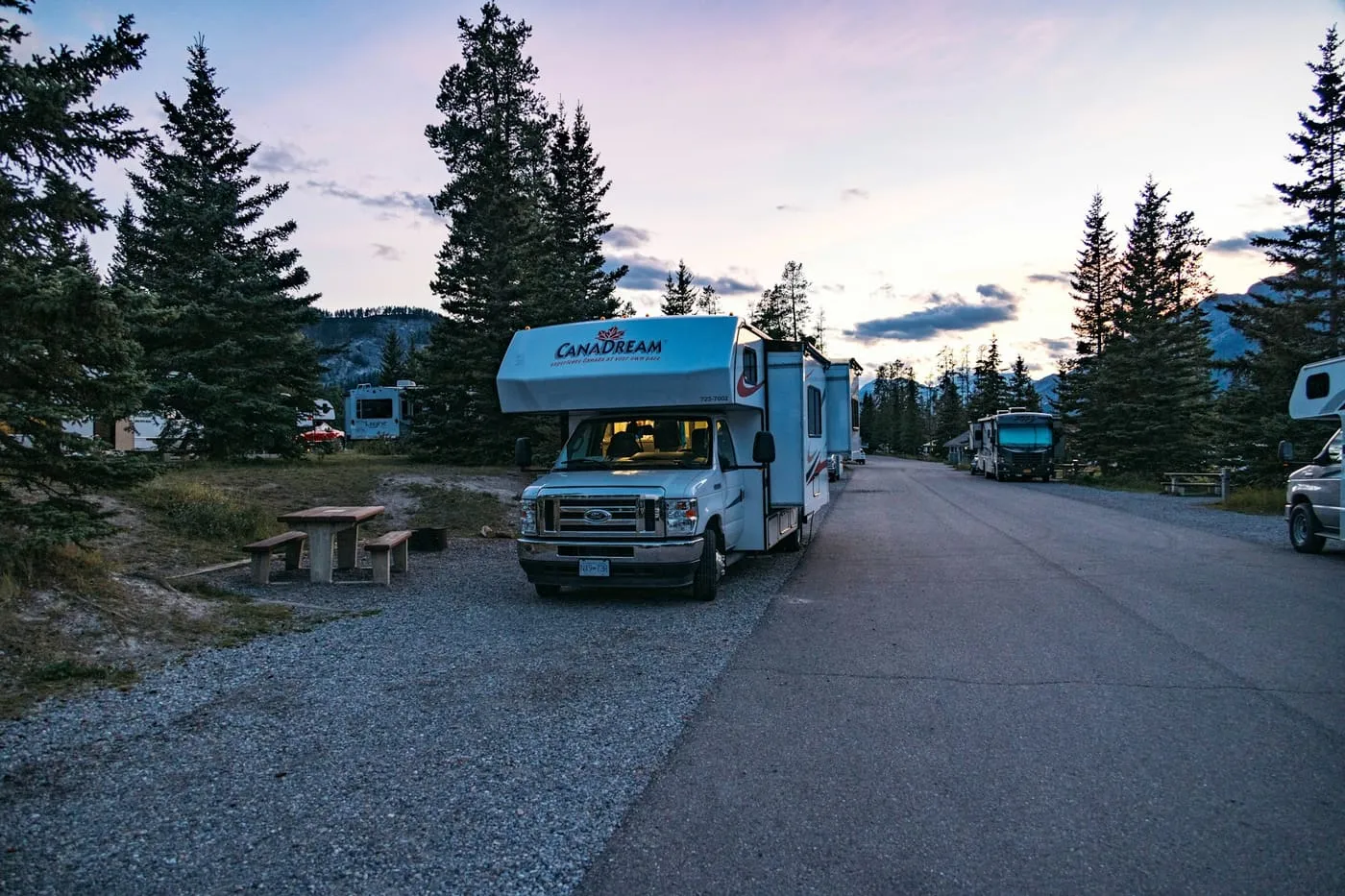 A motorhome parked on a gravel loop among spruce trees in a campground at twilight, in Banff, Alberta.