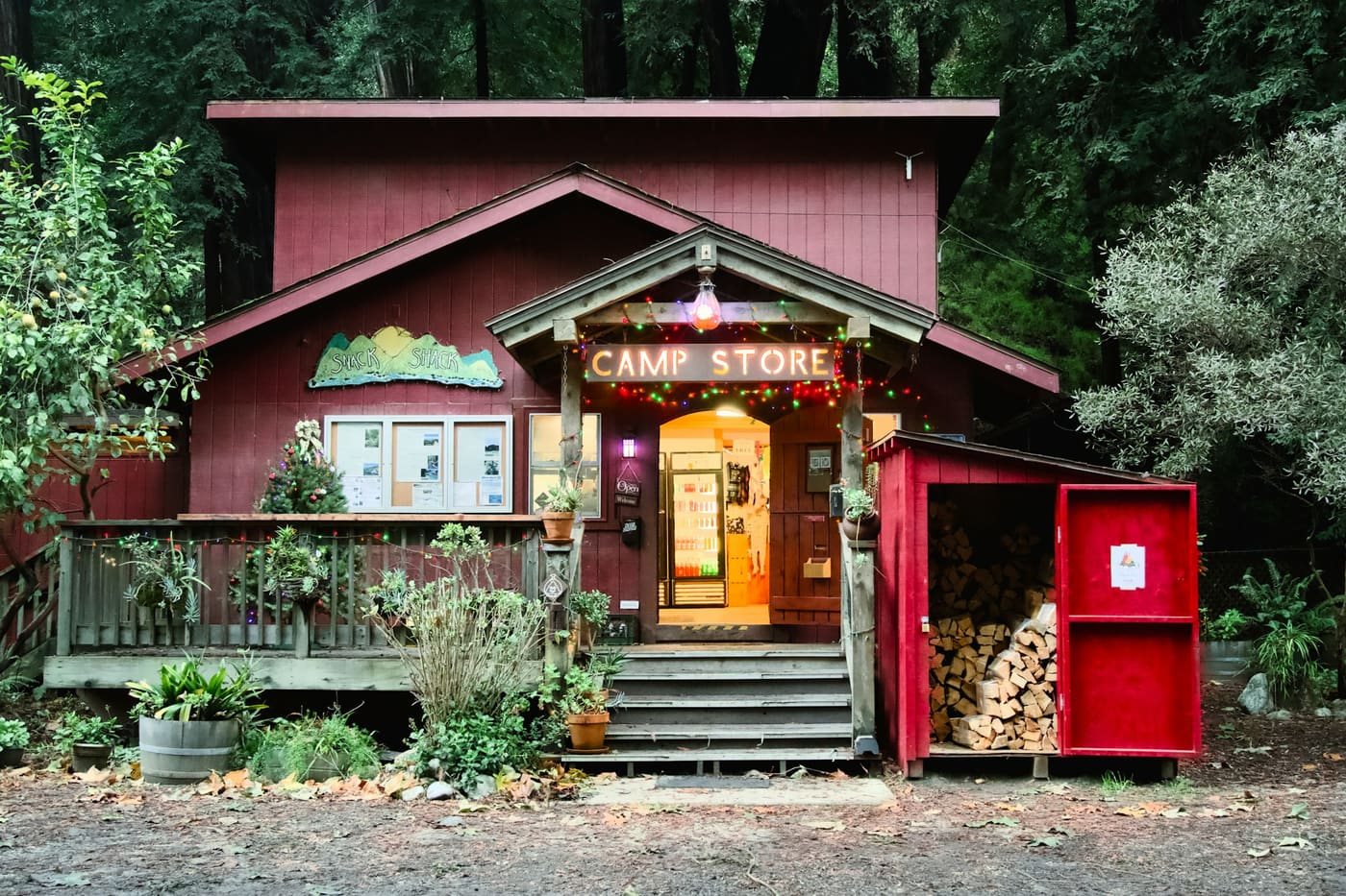 A red wooden camp store with a covered porch and a stack of firewood, with trees behind it.