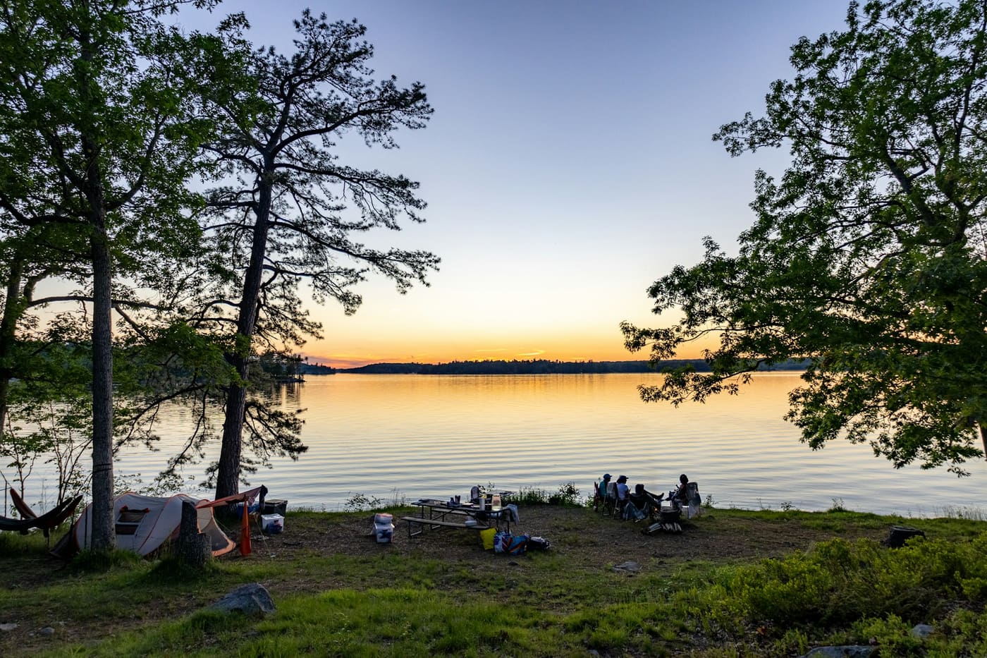People gathered on a grassy shore at sunset beside a calm lake, framed by tall trees.