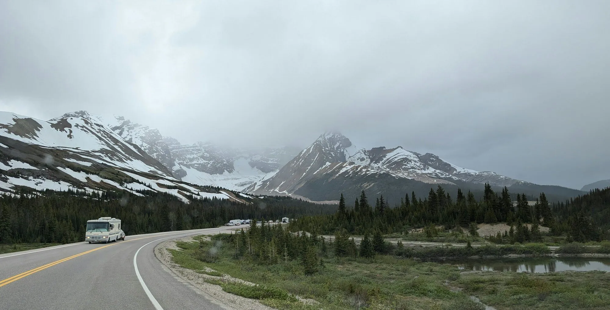 A motorhome on a curving two-lane highway below snow-streaked mountains and low cloud, in Alberta.