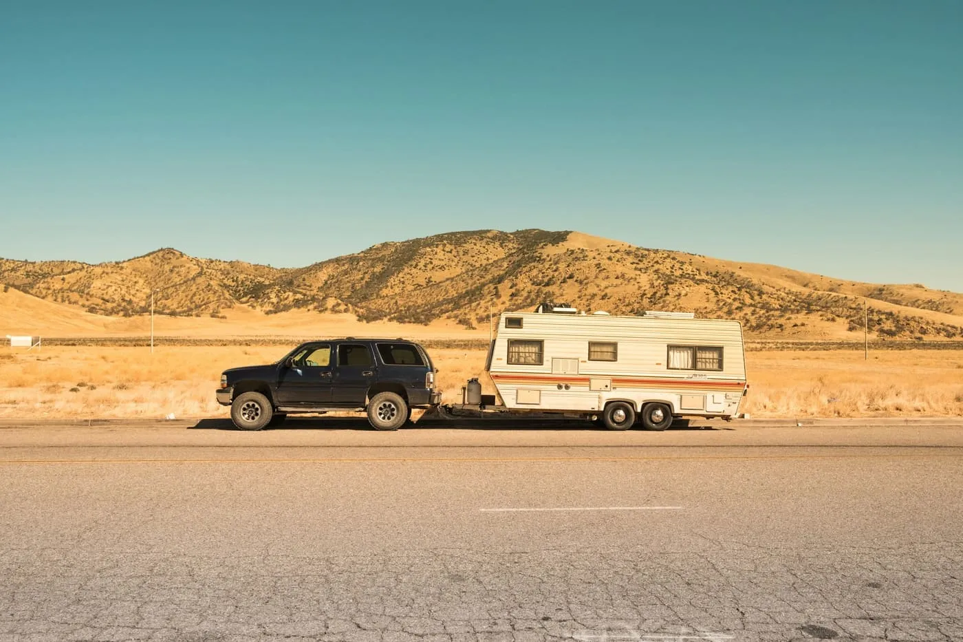 A black SUV towing a white travel trailer along a road below dry, golden hills.